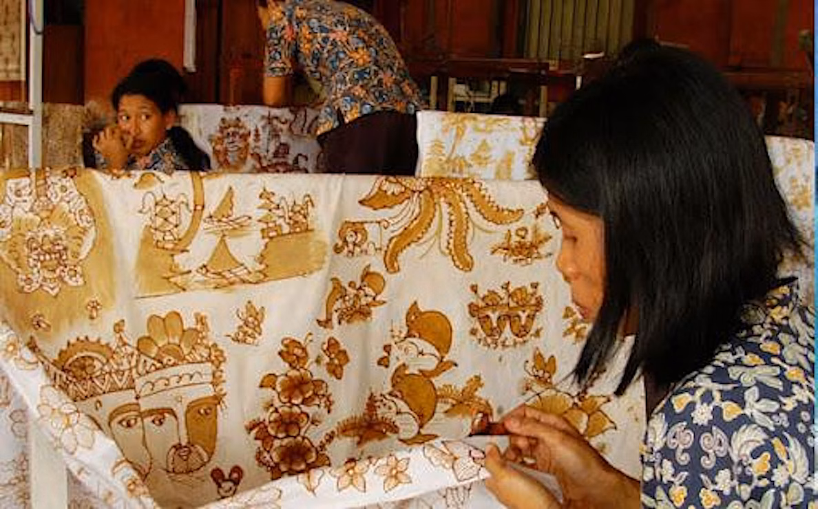 Three people work meticulously on traditional batik fabric, drawing intricate brown and gold patterns by hand. The scene suggests a workshop setting with focus on the detailed textile art.