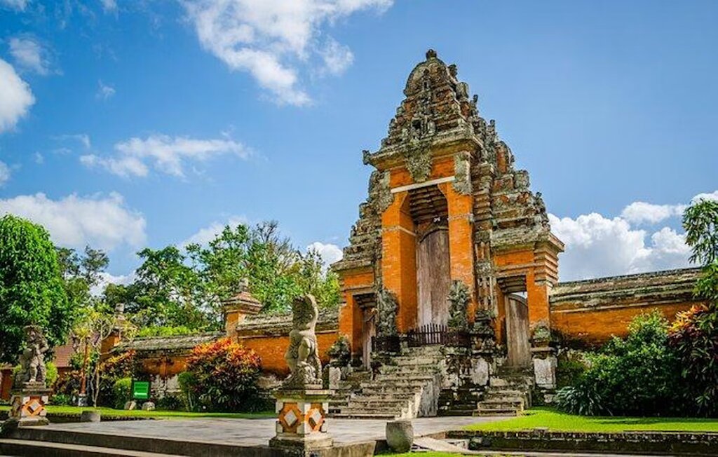 Traditional Balinese temple entrance with ornate stone carvings, stone statues, and orange brick, surrounded by lush greenery and blue sky with clouds.