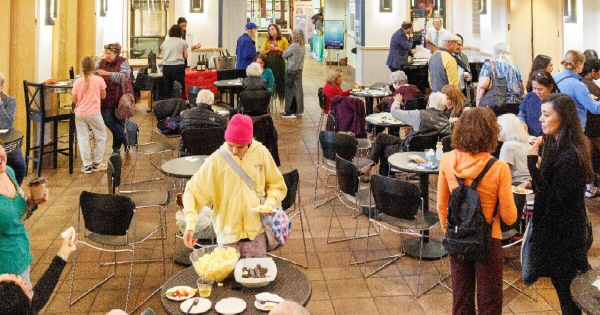 A lively indoor gathering with people of various ages sitting, eating, and socializing in a bright, spacious hall decorated with colorful banners and string lights.