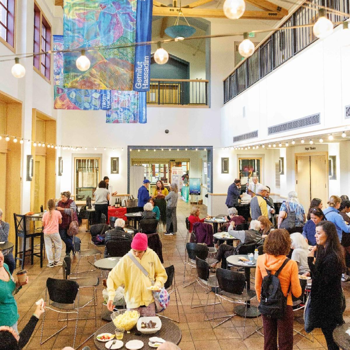 A lively indoor gathering with people of various ages sitting, eating, and socializing in a bright, spacious hall decorated with colorful banners and string lights.