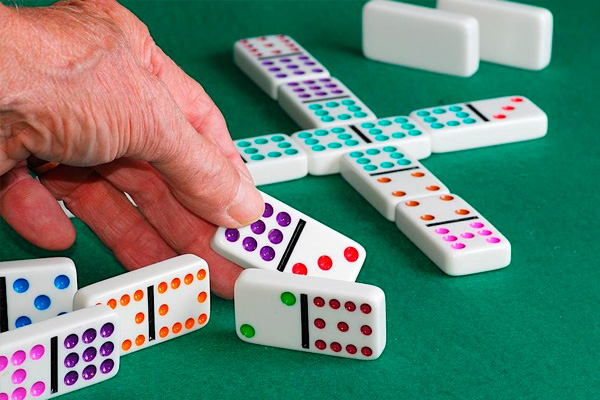 A hand is holding a domino tile over a green table at the Jewish Community Center, with several colorful dominoes in play and others scattered nearby.