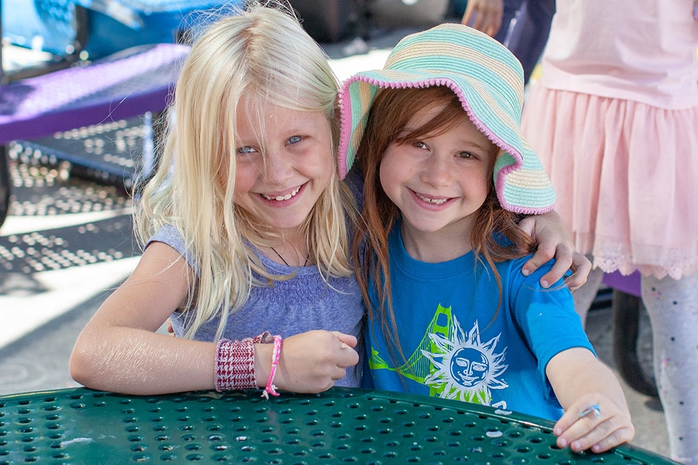 Two young girls smiling and hugging at an outdoor table. One has blonde hair and wears a lavender top; the other has brown hair, a colorful sun hat, and a blue shirt with a sun graphic. They appear happy and playful.