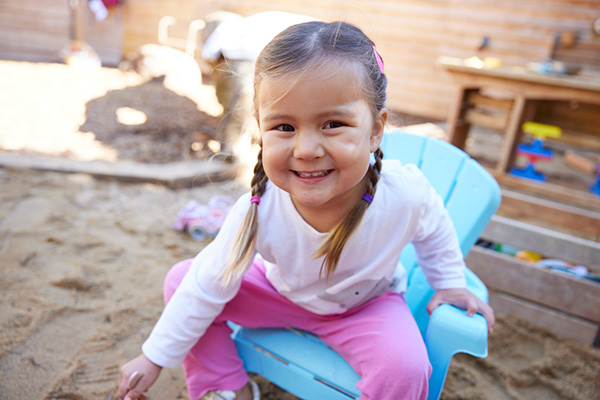 A young girl with brown hair in pigtails sits smiling on a blue chair in a sandy outdoor play area at a Jewish community center, wearing a white shirt and pink pants. Wooden toys and a sandbox are visible in the background.