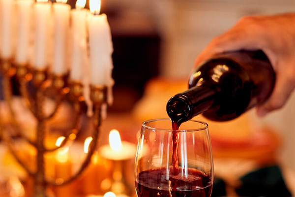 A hand pours red wine from a bottle into a glass beside a lit menorah, as several candles burn brightly in the background at a Jewish community center.