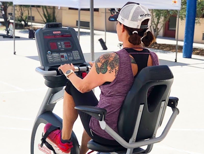 A woman in athletic wear and a cap rides a recumbent exercise bike outdoors, as if taking her gym routine beyond the gym reservation system. Her tattooed arm and bright sneakers stand out against sunny buildings and shaded areas.