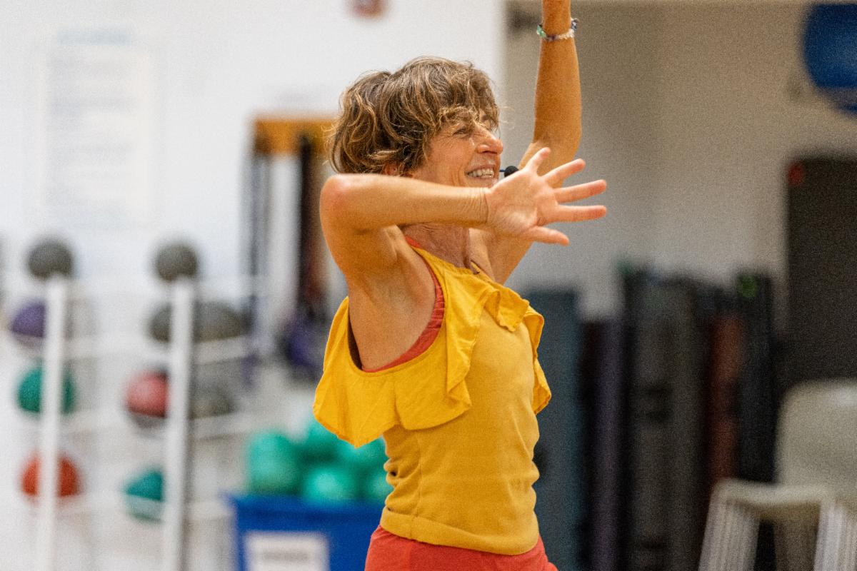 A woman wearing a yellow sleeveless top and red pants is smiling and raising her arms while exercising in a gym, with colorful exercise equipment visible in the background.