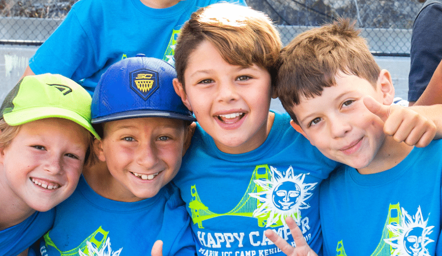 Four smiling boys wearing bright blue "Happy Camper" shirts and colorful caps pose together outdoors, giving thumbs up and hugging, with a fence and blurred background behind them.