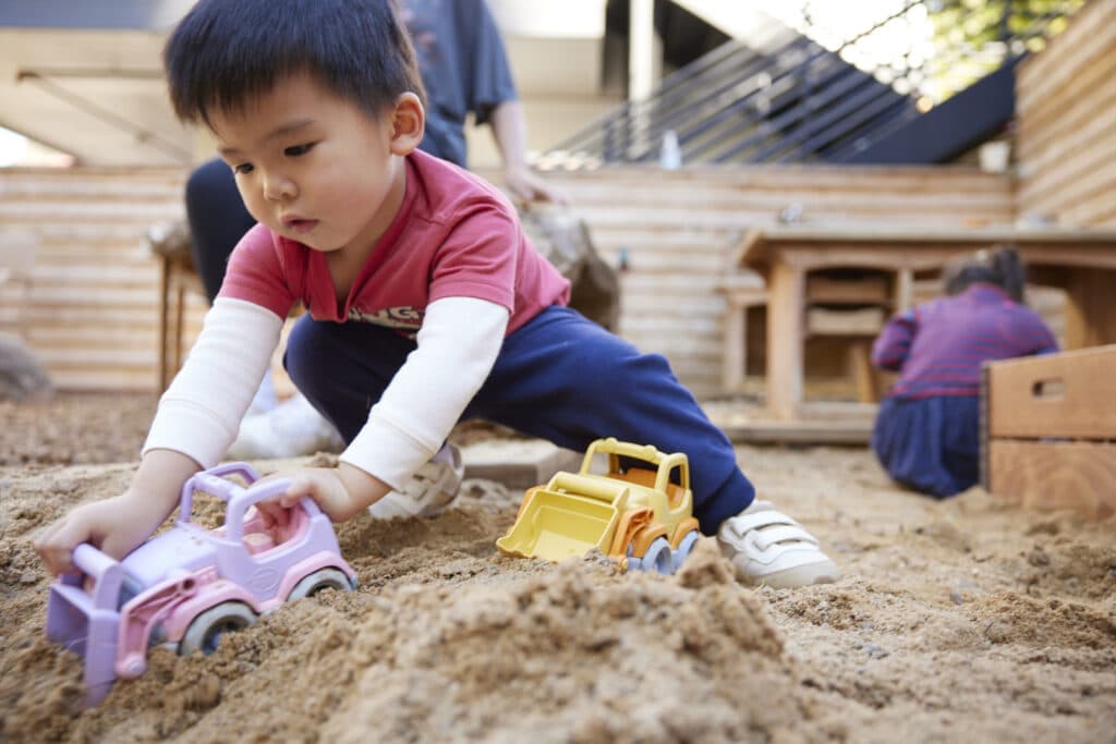 A young preschool child in a red shirt and white shoes plays with toy trucks in a sandy outdoor play area, focusing intently. Other children and adults are visible in the background.