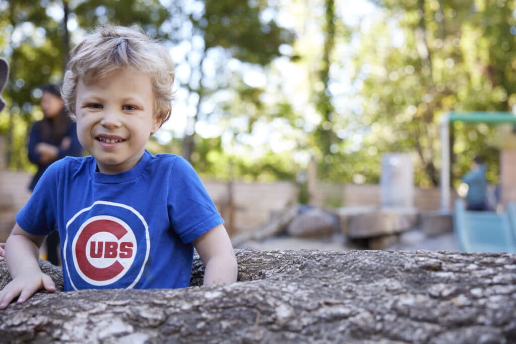 A preschool-aged child with blonde hair smiles while standing behind a large tree trunk, wearing a blue Chicago Cubs t-shirt. The background shows trees and a sunlit park area.