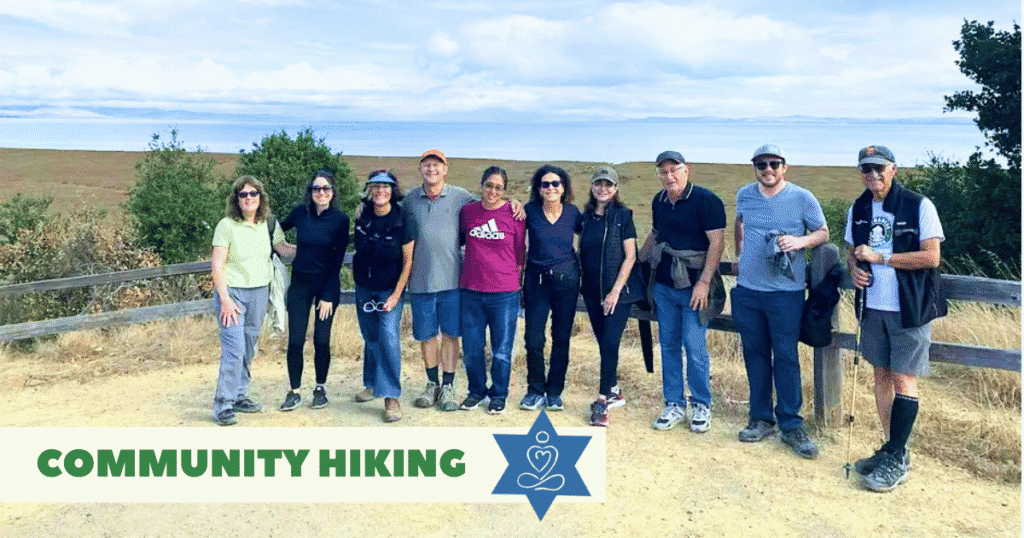 A group of ten people smiling and posing on a hiking trail with trees and open land behind them. A banner at the bottom reads "COMMUNITY HIKING" with a blue star and yoga design.