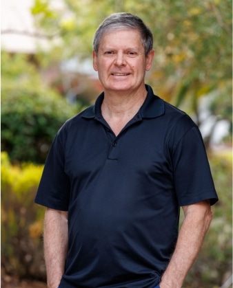 A man with short gray hair wearing a dark short-sleeve polo shirt stands outdoors, smiling, with greenery and trees blurred in the background—capturing the approachable confidence often seen in personal training professionals.