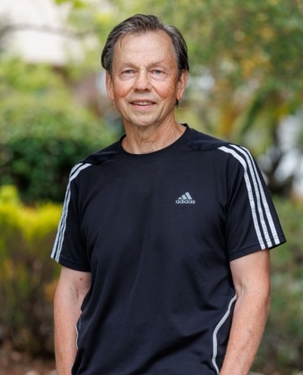 An older man wearing a black Adidas t-shirt stands outdoors with greenery in the background, smiling at the camera—radiating the confidence and energy that comes from personal training.