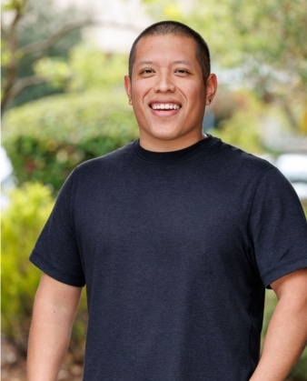 A smiling person with short hair and earrings stands outdoors, wearing a black t-shirt, radiating the confidence that comes from personal training, with greenery and blurred trees in the background.
