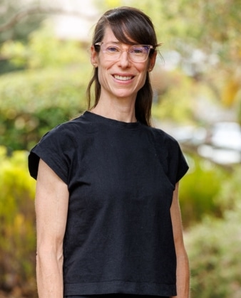 A woman with glasses and straight brown hair stands outdoors, smiling. She is wearing a black short-sleeved top, exuding a friendly and approachable presence often seen in personal training professionals. The background is blurred greenery.