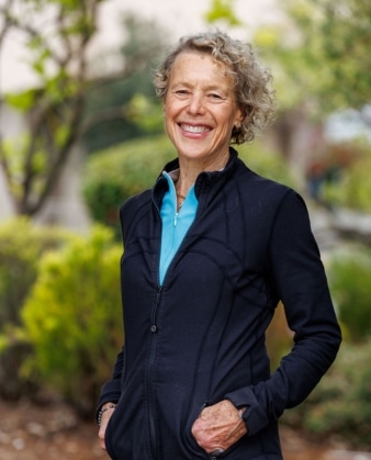 Smiling older woman with curly gray hair stands outdoors, wearing a blue shirt and black zip-up jacket, with hands in her jacket pockets. Green foliage and trees appear in the blurred background, suggesting she might be enjoying a break during her personal training session.