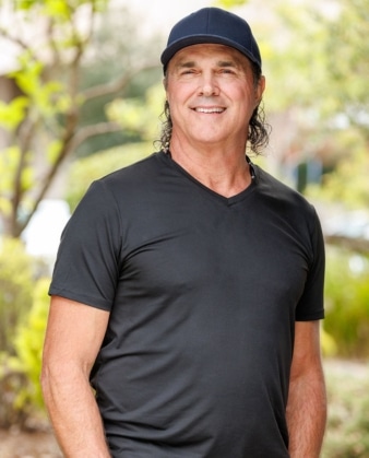 A middle-aged man with long dark hair, wearing a black V-neck T-shirt and a navy blue cap, smiles outdoors in a leafy setting, embodying the approachable energy of someone dedicated to personal training.