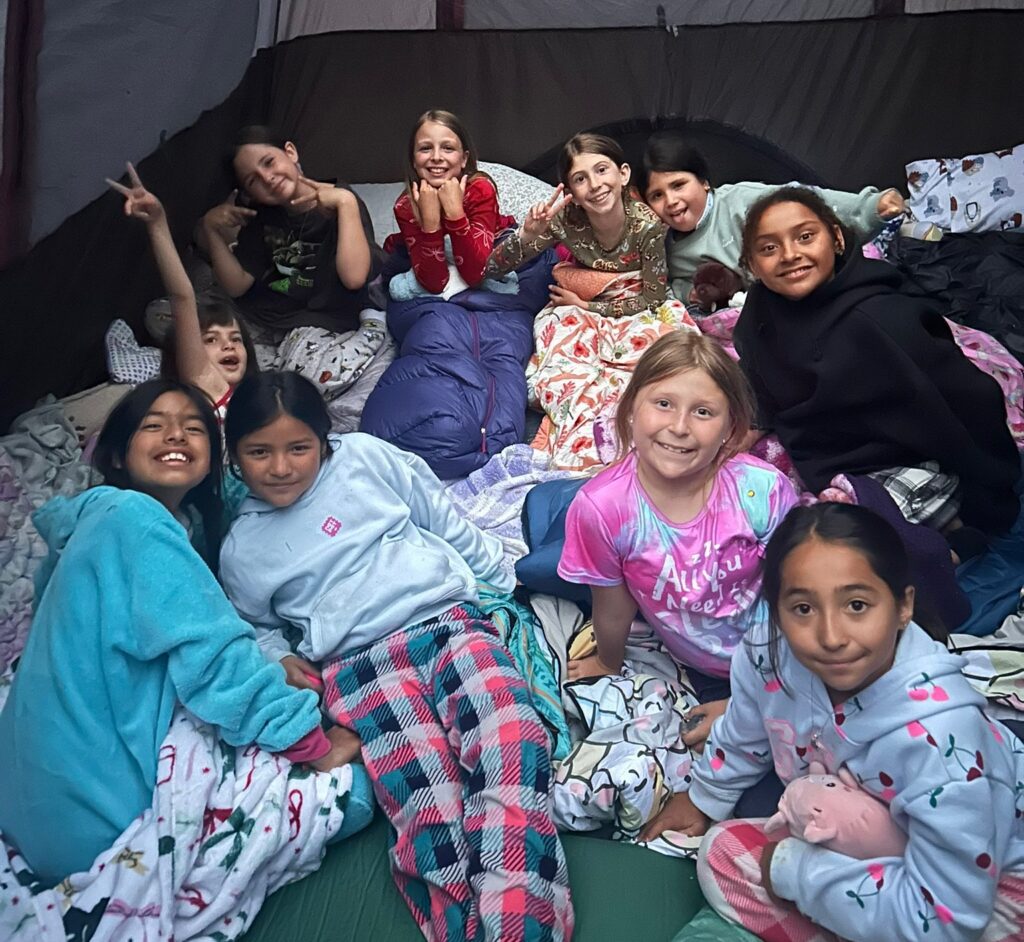 Eleven girls in pajamas smile and pose inside a tent, sitting and lying on sleeping bags and blankets, suggesting a fun sleepover or camping trip.