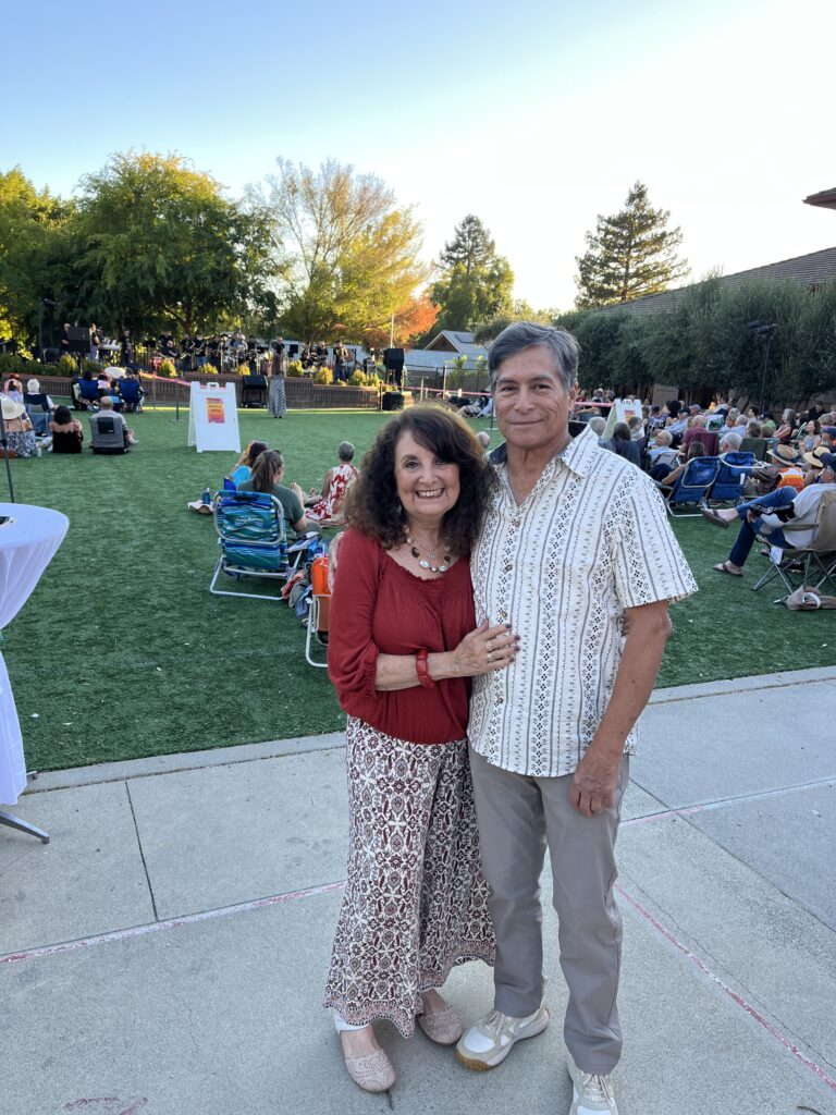A smiling couple stands together at an outdoor event on a grassy area, with people seated behind them and musicians performing on a stage in the background during daylight.