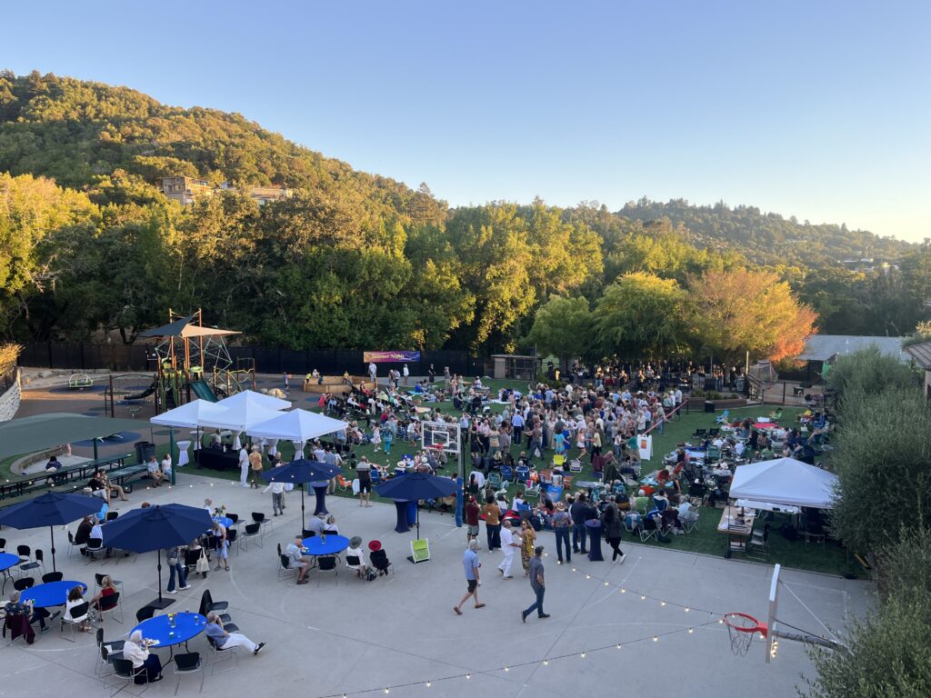 A large outdoor gathering with people seated at tables and mingling on a lawn, surrounded by trees and hills under a clear sky at sunset. Tents, string lights, and a playground are visible in the background.