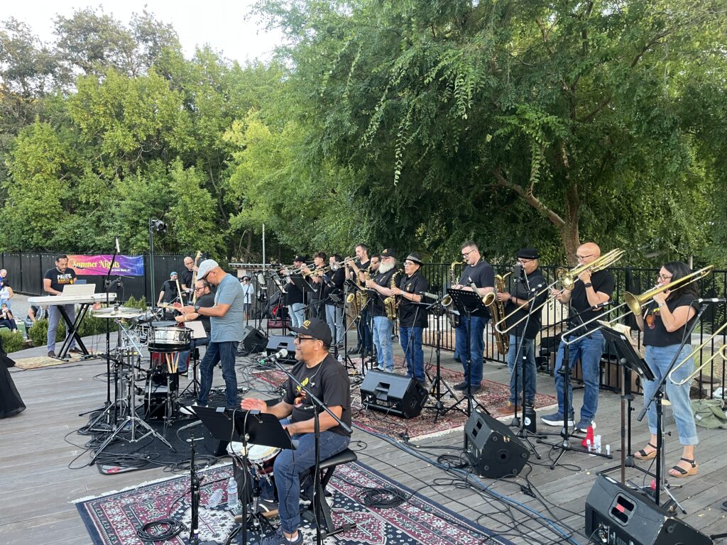 A large band performs outdoors on a wooden stage with microphones, keyboards, brass instruments, and drums, surrounded by trees and greenery. The musicians wear black shirts and play to an unseen audience.