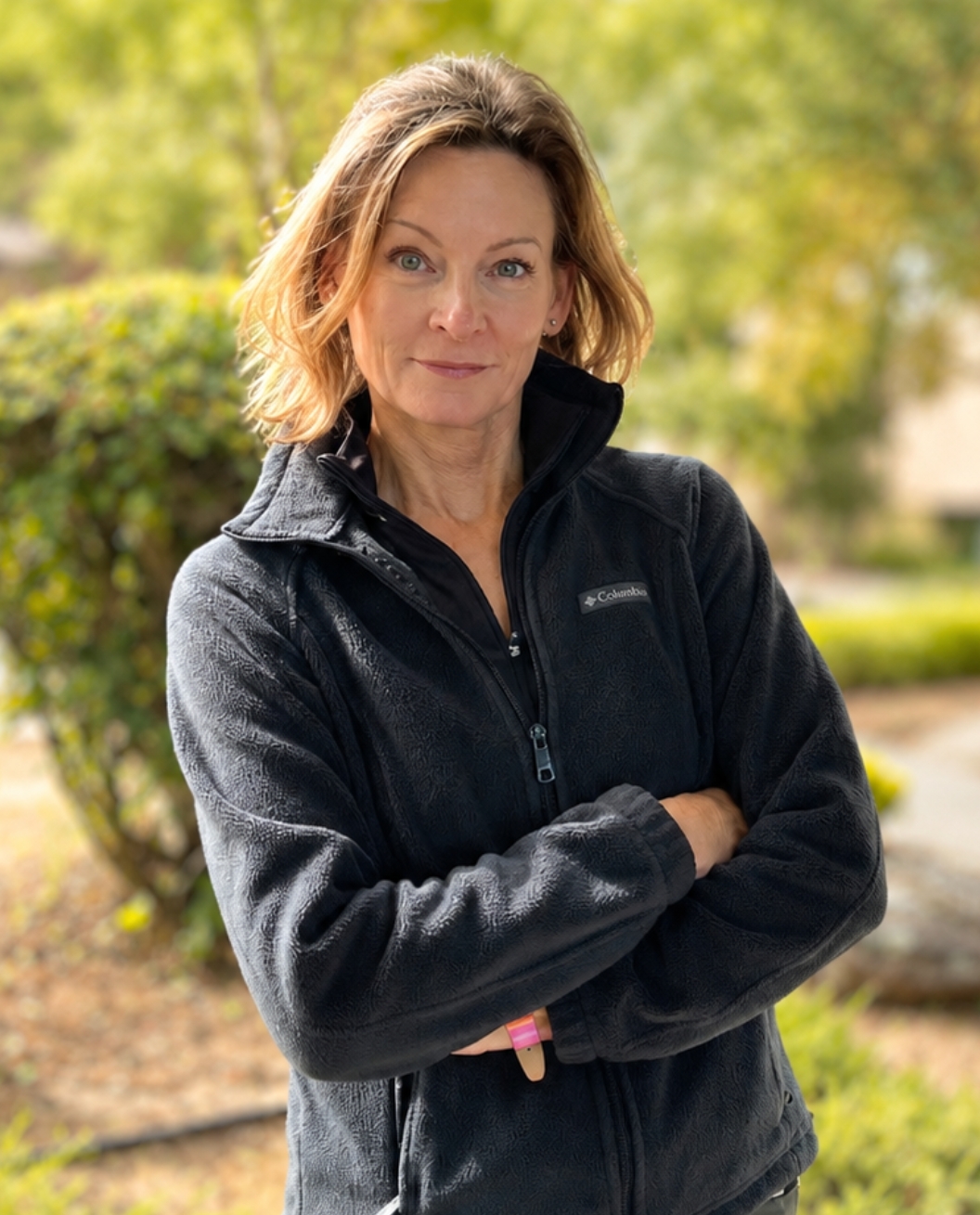A woman with short, light brown hair stands outdoors with arms crossed, wearing a black Columbia fleece jacket—her confident stance reflecting a dedication to personal training. Greenery and blurred trees are in the background.
