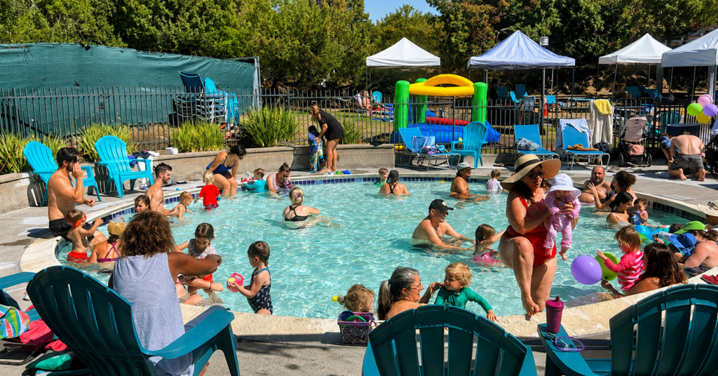 Children and adults play and relax in a small, crowded outdoor pool on a sunny day. Some sit on pool chairs, while others wade in the water. Tents, a bounce house, and trees are visible in the background.