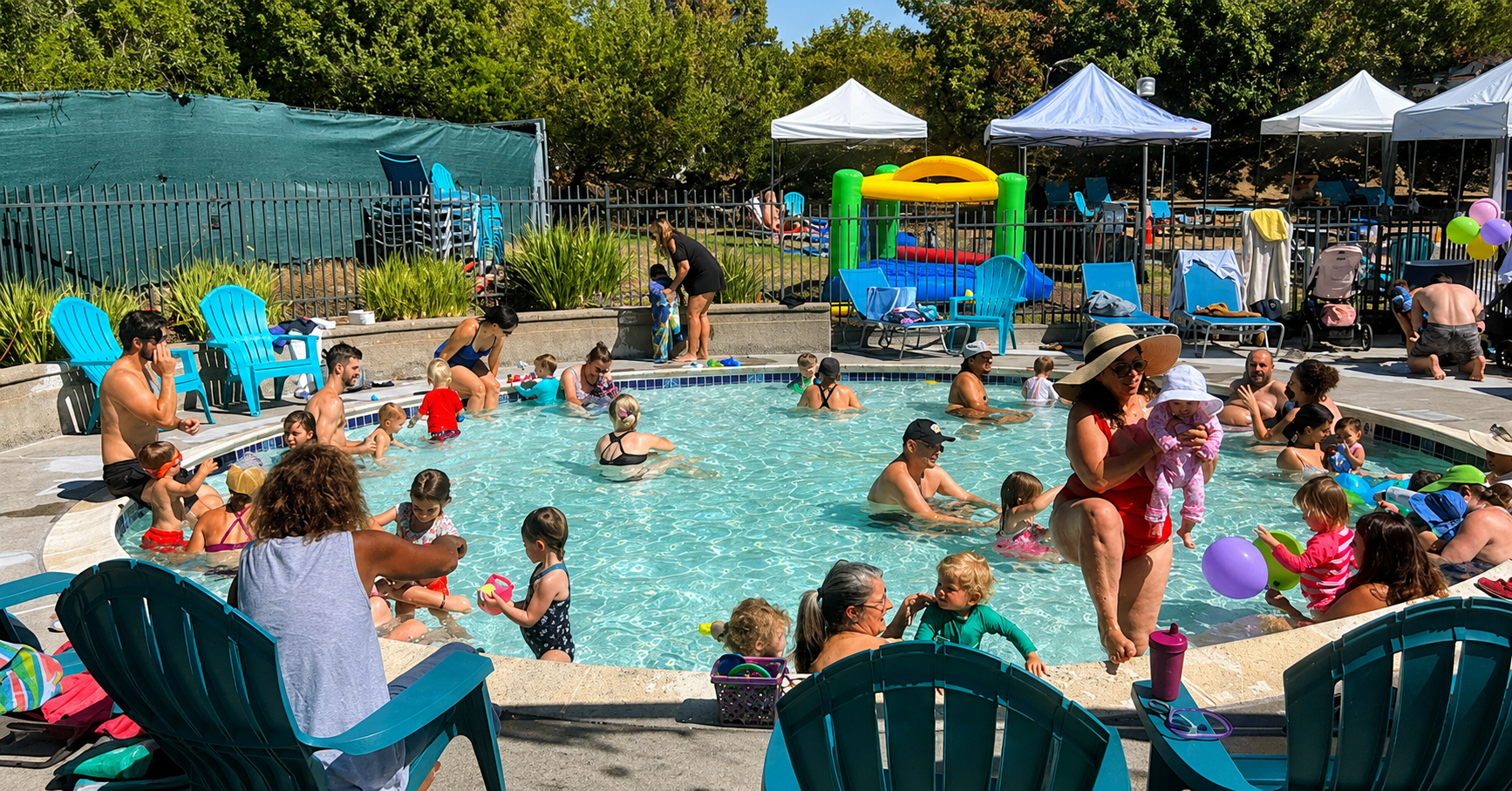 Children and adults play and relax in a small, crowded outdoor pool on a sunny day. Some sit on pool chairs, while others wade in the water. Tents, a bounce house, and trees are visible in the background.