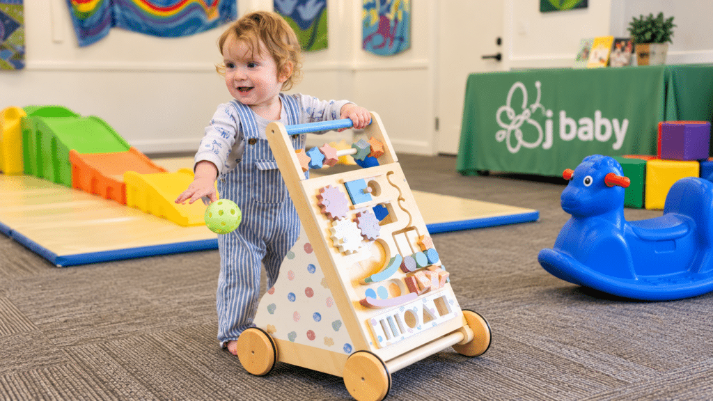 A toddler with curly hair pushes a colorful wooden walker while holding a green ball in a playroom with toys, a rocking horse, and a green table with “baby” written on it.