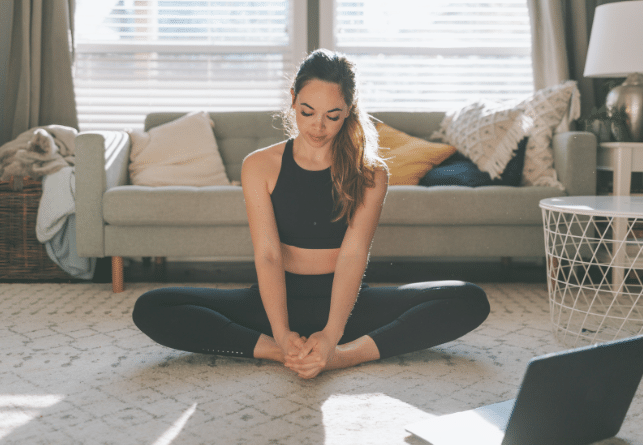 Woman in black workout clothes sits on a living room floor in a butterfly stretch. Sunlight streams through the windows behind her, and a laptop is open on the rug in front of her.