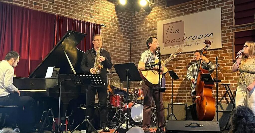 Five musicians perform on stage: a pianist, saxophonist, guitarist, upright bassist, and singer. A brick wall and a sign reading “the backroom” are in the background. Red curtains and stage lights are visible.