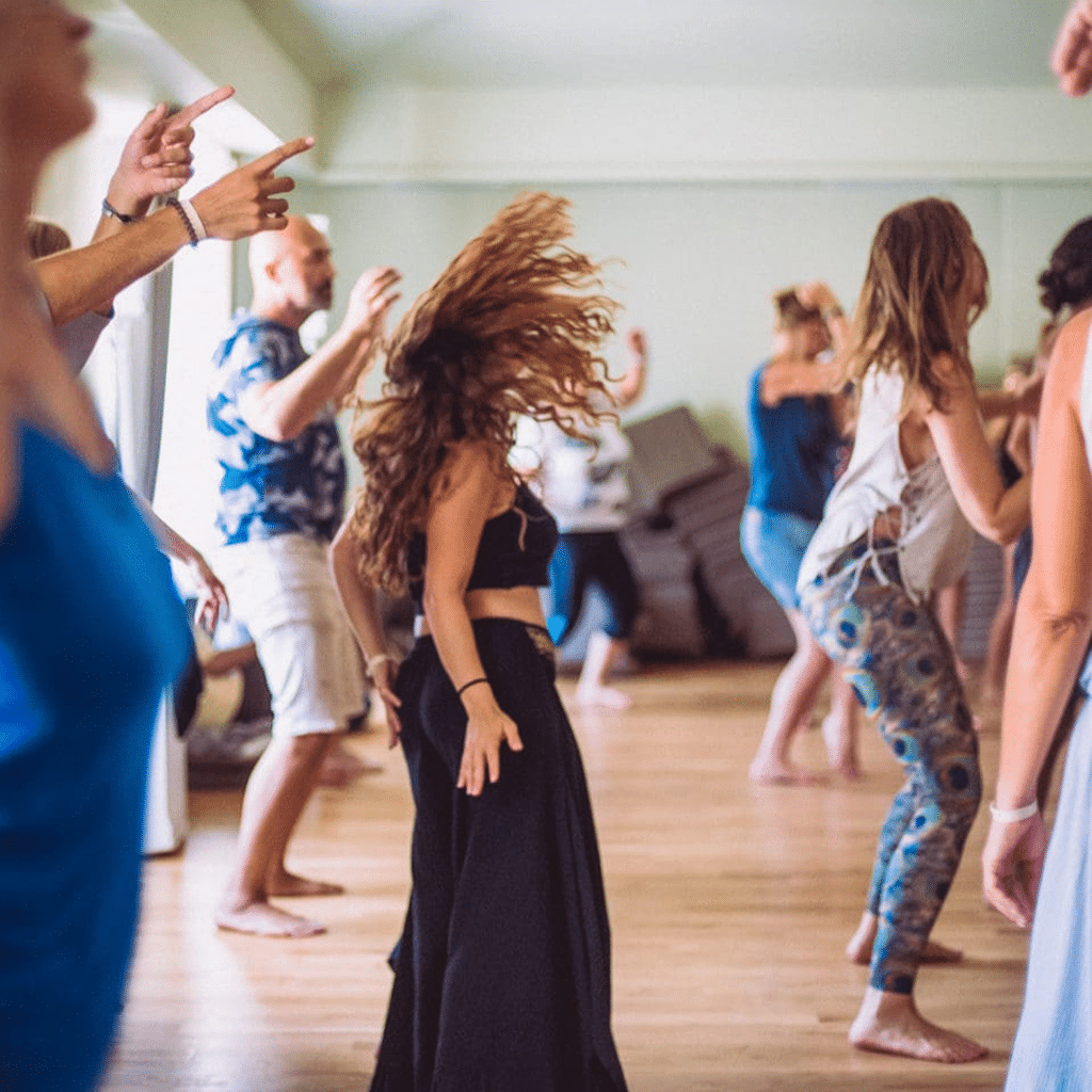 People dancing barefoot in a bright room, with one woman in the foreground flipping her long hair. Others move energetically, wearing casual, colorful clothing. The atmosphere is lively and expressive.