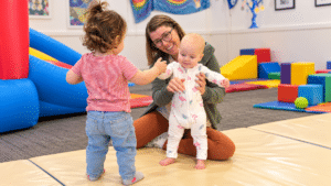 A smiling adult helps a baby in pajamas stand on a play mat while another toddler in jeans and a striped shirt faces them in a colorful indoor play area.