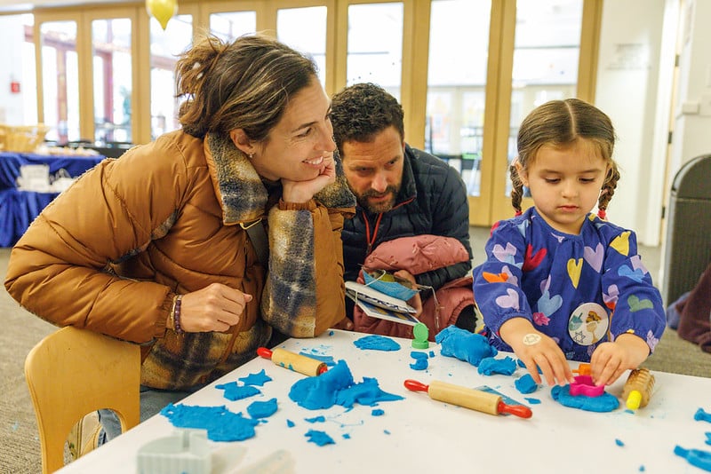 A young girl sits at a table playing with blue modeling clay, watched by a smiling woman and a man.