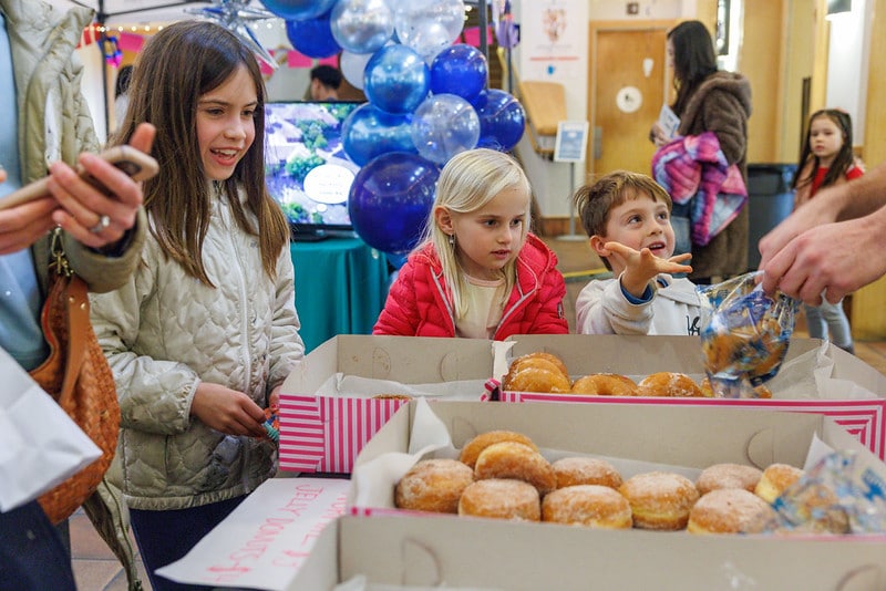 A boy reaches for a donut from a plastic bag held by an adult as two young girls look on, with boxes of jelly donuts in the foreground.