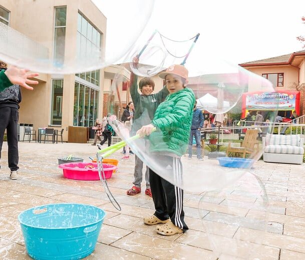 A child in a green jacket creates large bubbles with a wand in a plaza with other children and bubble solution tubs.