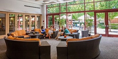 A spacious lounge with large windows and glass doors, featuring several people sitting and chatting on orange cushioned sofas arranged in a semi-circle. Outside, people are visible on a sunny patio with tables and umbrellas.
