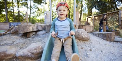 A young preschool boy wearing a red cap slides down an outdoor playground slide, smiling happily. The playground is surrounded by rocks, logs, and trees, with a woman sitting in the background.