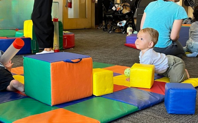 Two toddlers play on colorful foam mats with large foam blocks. One child crawls and looks up, while another lies down holding a tube. An adult and another child sit in the background.