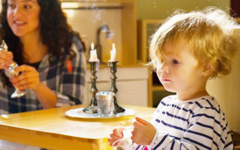 A young child in a striped shirt stands near a table with lit candles, while an adult woman smiles in the background, holding a glass. Warm sunlight fills the cozy room.