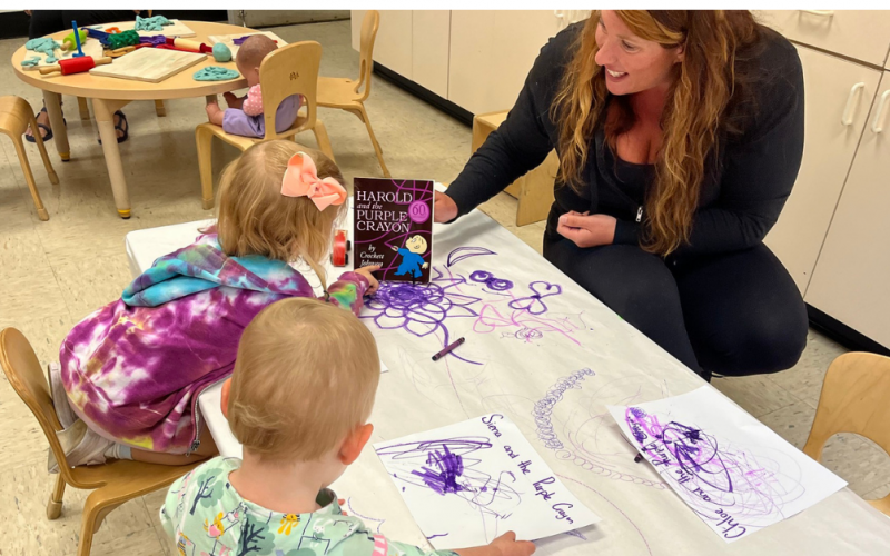 A woman smiles while two young children draw with purple markers at a table. A book, “Harold and the Purple Crayon,” is propped up nearby. The scene takes place in a classroom with toys and small chairs.