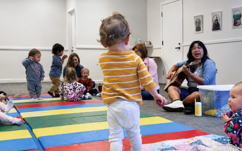 A group of toddlers sit and play on colorful mats in a classroom, while adults supervise. One adult plays a guitar and sings, engaging the children. The room has light walls and a cheerful atmosphere.
