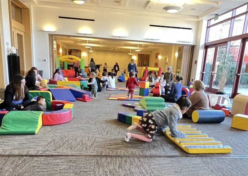 Children and adults play in a large indoor preschool space filled with colorful soft play equipment, including mats, blocks, and tunnels. Natural light streams in through large windows on the right.