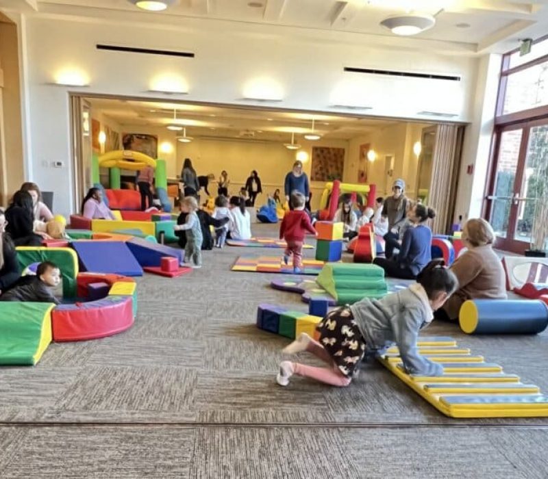 Children and adults play in a large indoor preschool space filled with colorful soft play equipment, including mats, blocks, and tunnels. Natural light streams in through large windows on the right.